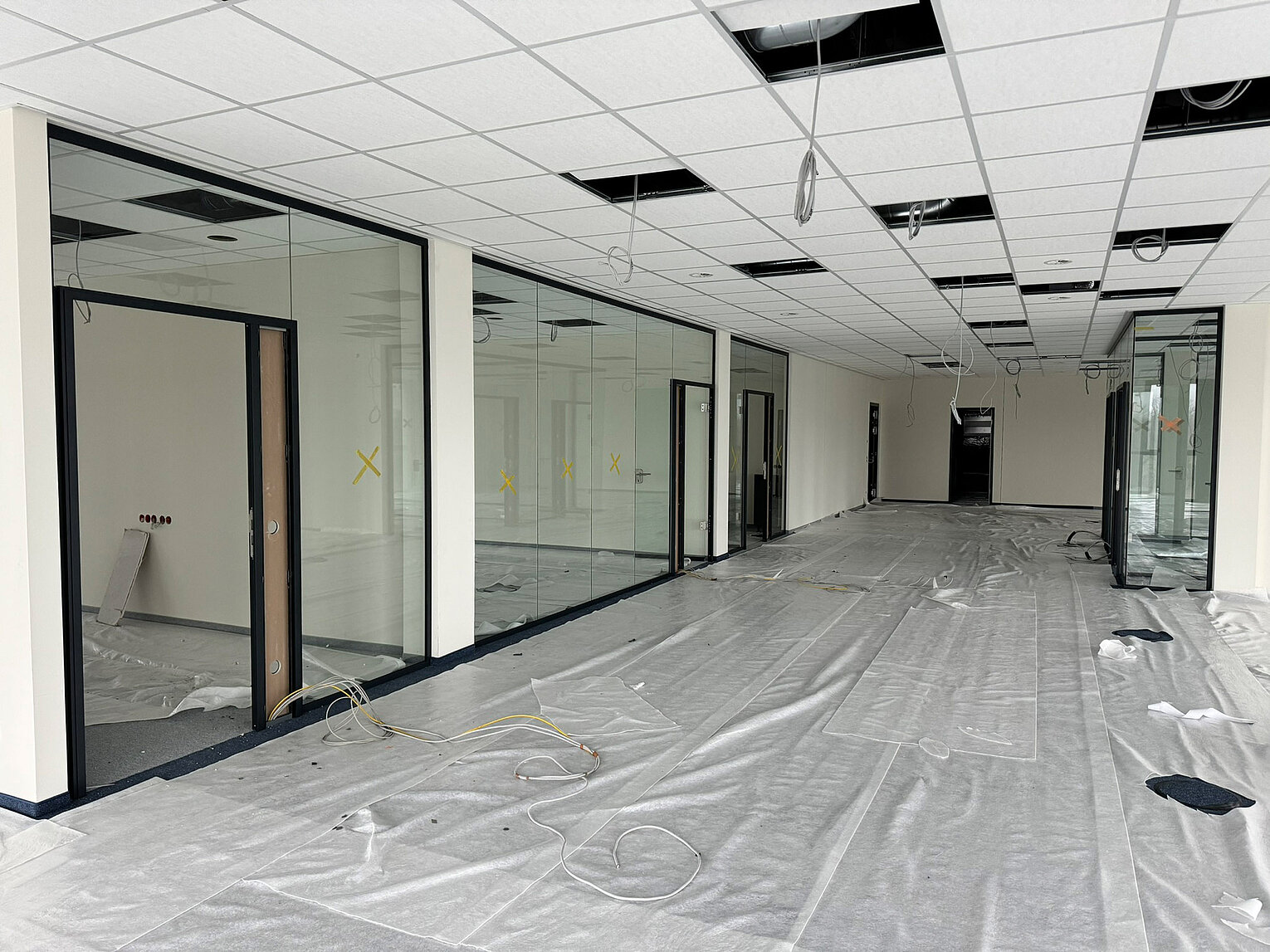 Amann Girbach Großer Büroraum im Rohbau mit abgehängter Decke, Glaswänden und provisorisch abgedechtem Boden, Kabeln liegen lose herum, Raum ist unmöbliert und wird renoviert.